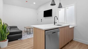 A modern kitchen with a dishwasher, a small island, pendant lights, a flat-screen TV, and a dark-colored sofa in a bright room with wooden flooring. - "Avanta" with free parking near UofC (Calgary)