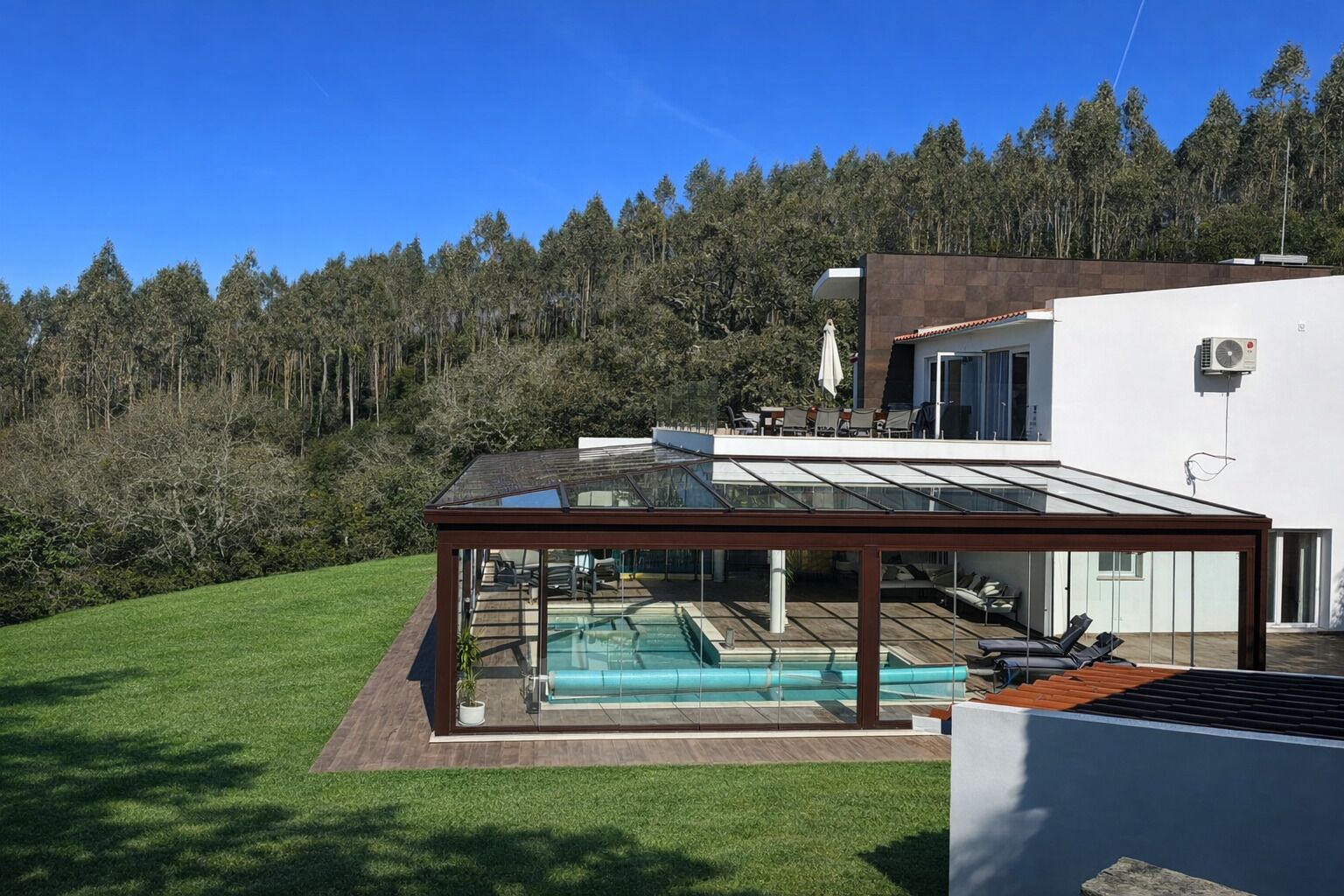Una casa moderna con piscina cubierta por vidrio, una terraza de madera y vista a una colina boscosa.