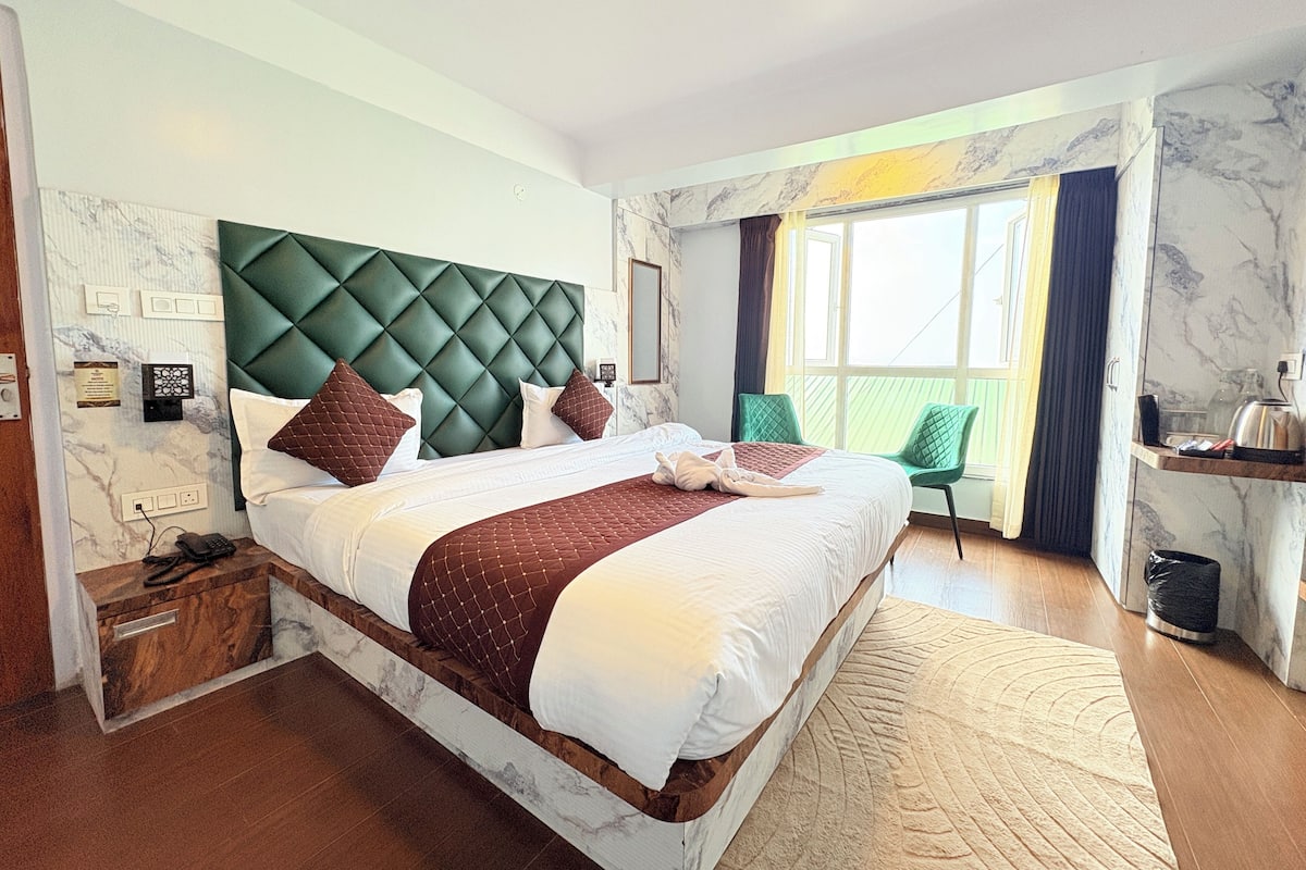 Quarto de hotel com uma cama grande, cabeceira estofada verde, parede com padrão marmorizado, janela com cortinas, pequena escrivaninha com chaleira e uma lixeira.