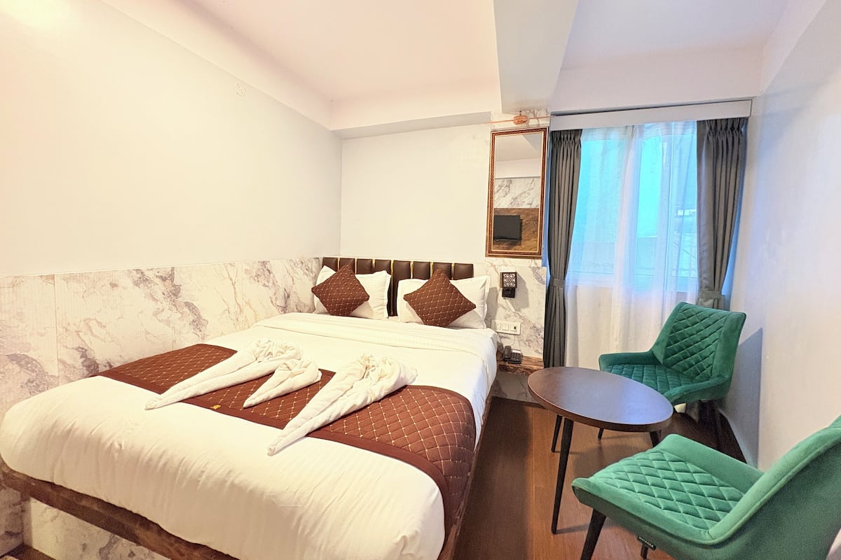 Quarto de hotel com cama, uma pequena mesa, duas cadeiras, espelho e janela com cortinas.