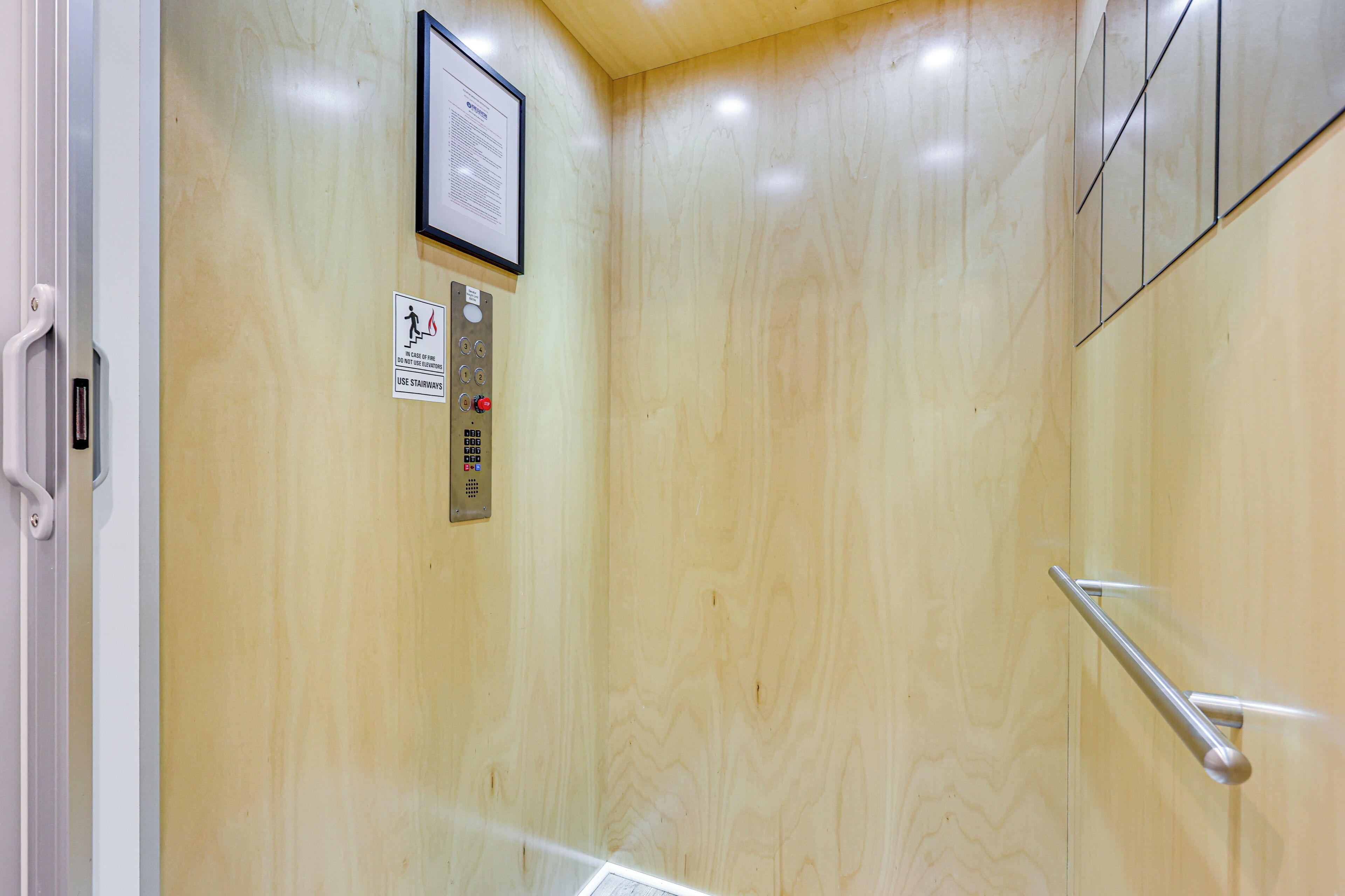 An elevator interior with a wooden wall, a framed notice, and a control panel with a red emergency button.