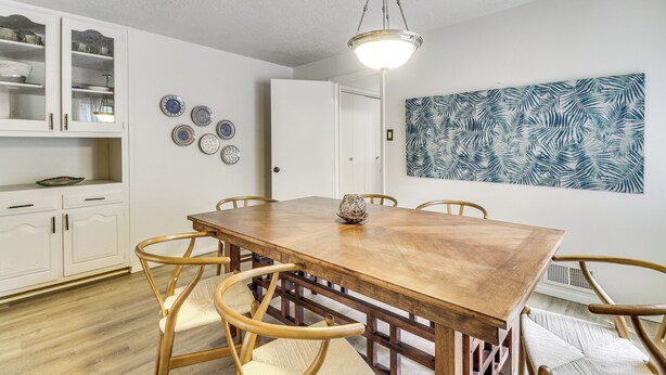 Un coin repas avec une table en bois et des chaises en osier, un buffet blanc aux portes en verre et un mur orné d’une œuvre d’art à motifs végétaux.