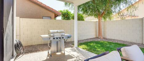 A covered patio area with a grill, a chair, and a small lawn.
