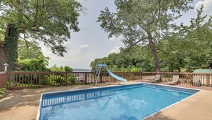 A rectangular swimming pool with a blue slide and a ladder, surrounded by a wooden deck and trees. - Hot Tub, Pool, Dock: 2 Cabins Lakeview Grand Lake (Grove)
