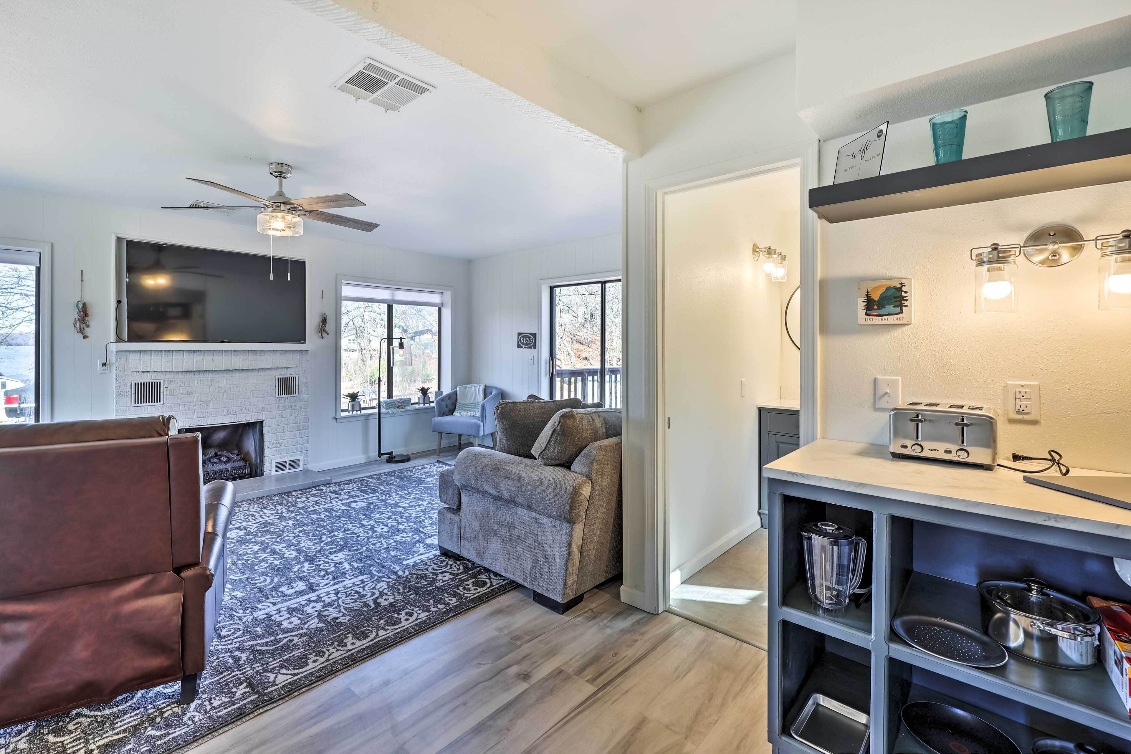 A modern living room with a fireplace, a ceiling fan, and a kitchen area with a toaster and shelves.