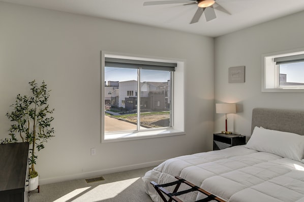 A bedroom with a bed, a nightstand, a lamp, a potted plant, and a window with a view of buildings. - Rooftop Oasis: Downtown Des Moines Townhome (Des Moines)