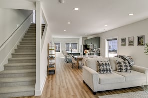 Um interior moderno com escada, sala de estar com sofá e almofadas, área de jantar com mesa e cadeiras, e cozinha com armários e eletrodomésticos.