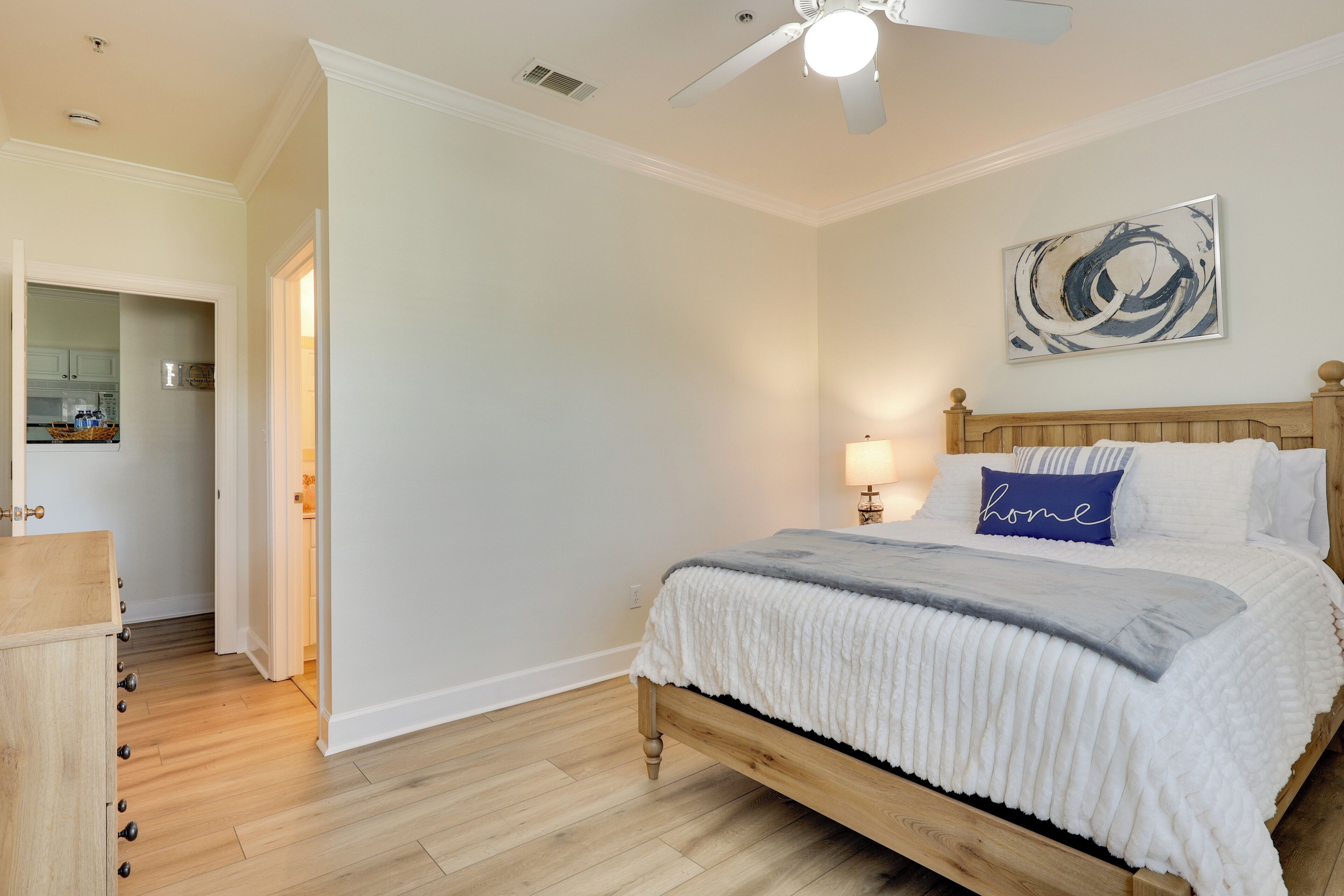 A bedroom with a wooden bed, a ceiling fan, a wall-mounted abstract painting, and a kitchen visible through an open door.