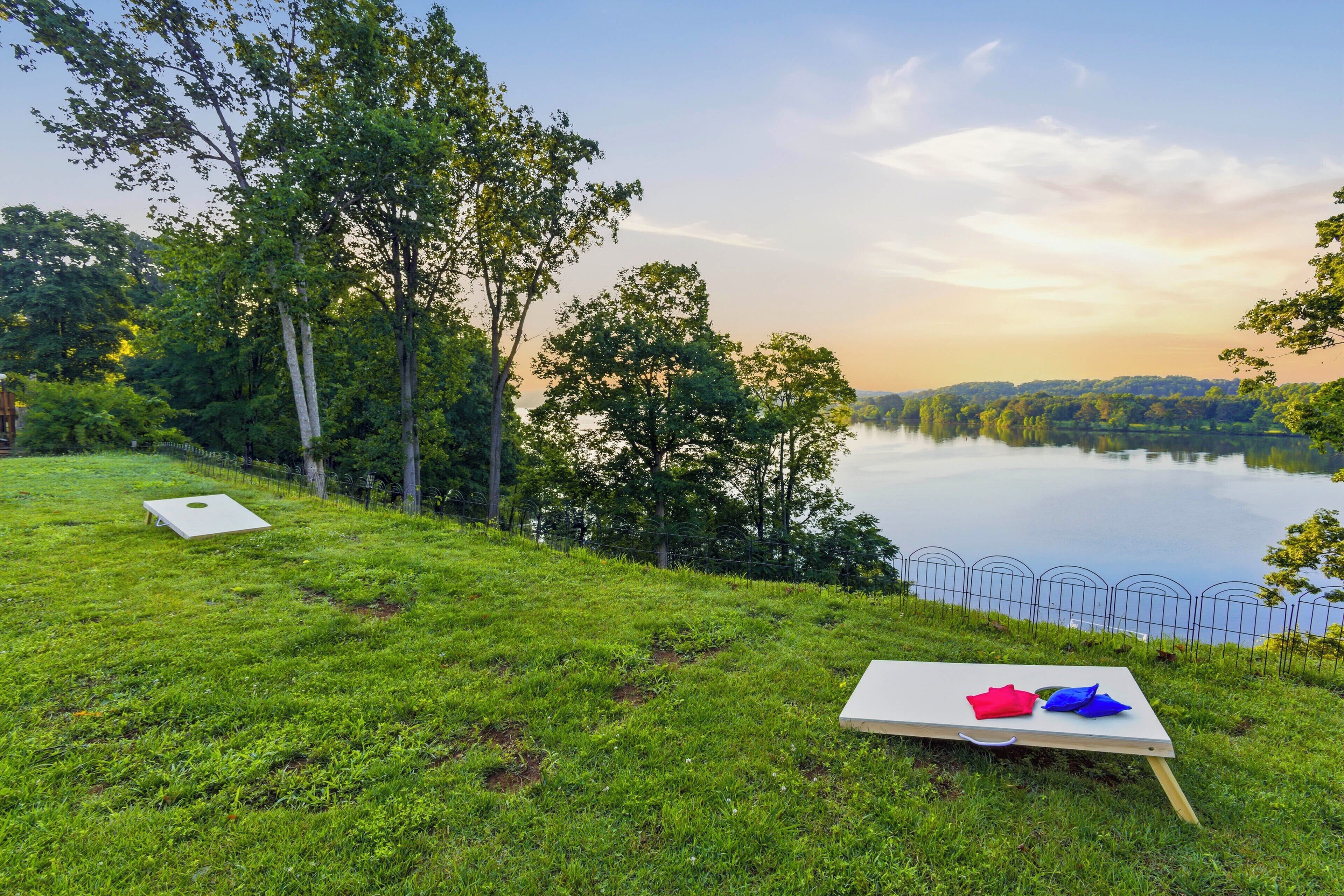 A grassy area with a picnic table and a white object, overlooking a calm lake with trees and a fence.