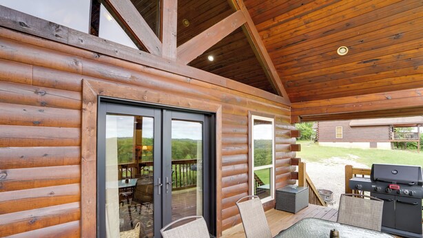 Eine Blockhütte mit Glastür, die auf eine Terrasse im Freien führt, einem Grill und einem Tisch mit Stühlen.
