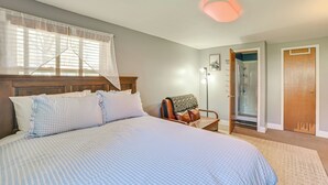 A bedroom with a large bed, a wooden headboard, a window with sheer curtains, a nightstand, a chair with a blanket, and a door leading to a bathroom. - Walk to the Beach! Boyne City Escape w/ Lake Views (Boyne City)