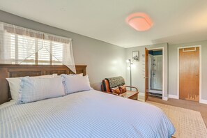 Une chambre avec un grand lit, une tête de lit en bois, une fenêtre avec des rideaux légers, une table de chevet, une chaise recouverte d’une couverture et une porte donnant sur une salle de bain.