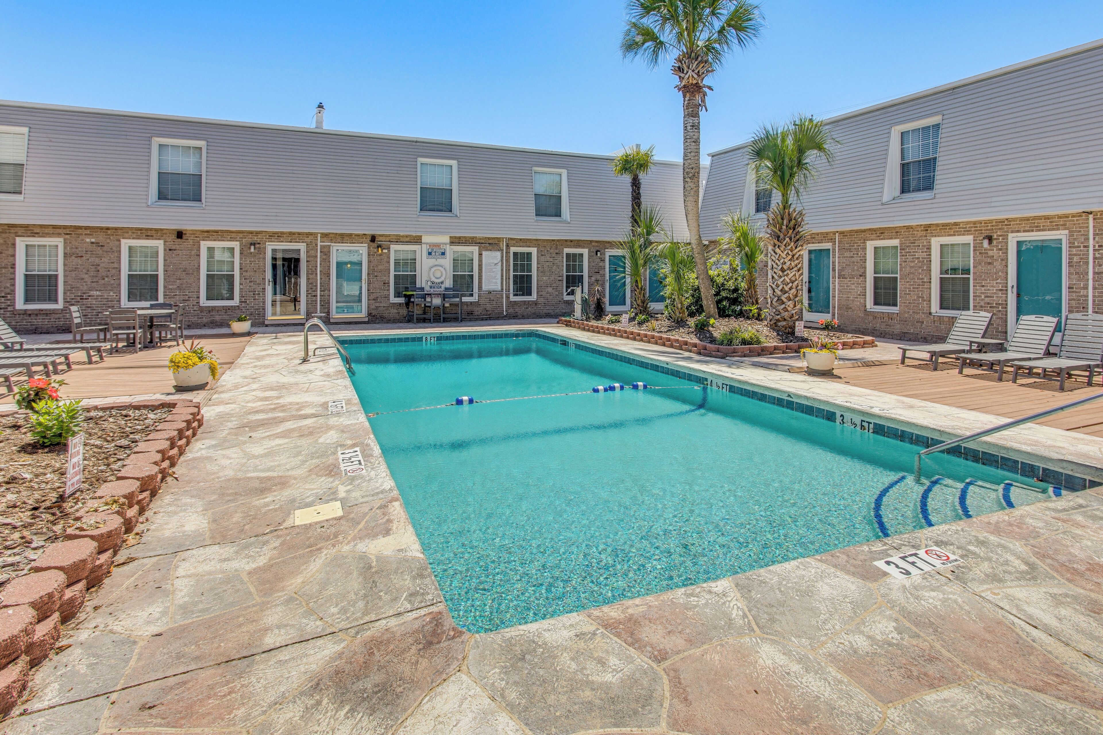 A rectangular pool with clear blue water, surrounded by a stone deck, and adjacent to a building with brick and siding exteriors.