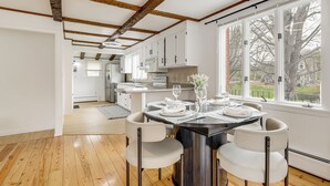 Una cocina moderna con armarios blancos, una zona de comedor con una mesa oscura y un ventanal con vistas a árboles.