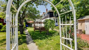Un chemin menant à une arche blanche, entourée de verdure, avec une maison en arrière-plan.