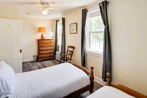 Ein Schlafzimmer mit zwei Einzelbetten, einem Schminktisch, einem Sessel, einem Heizkörper und einem Fenster mit Vorhängen.