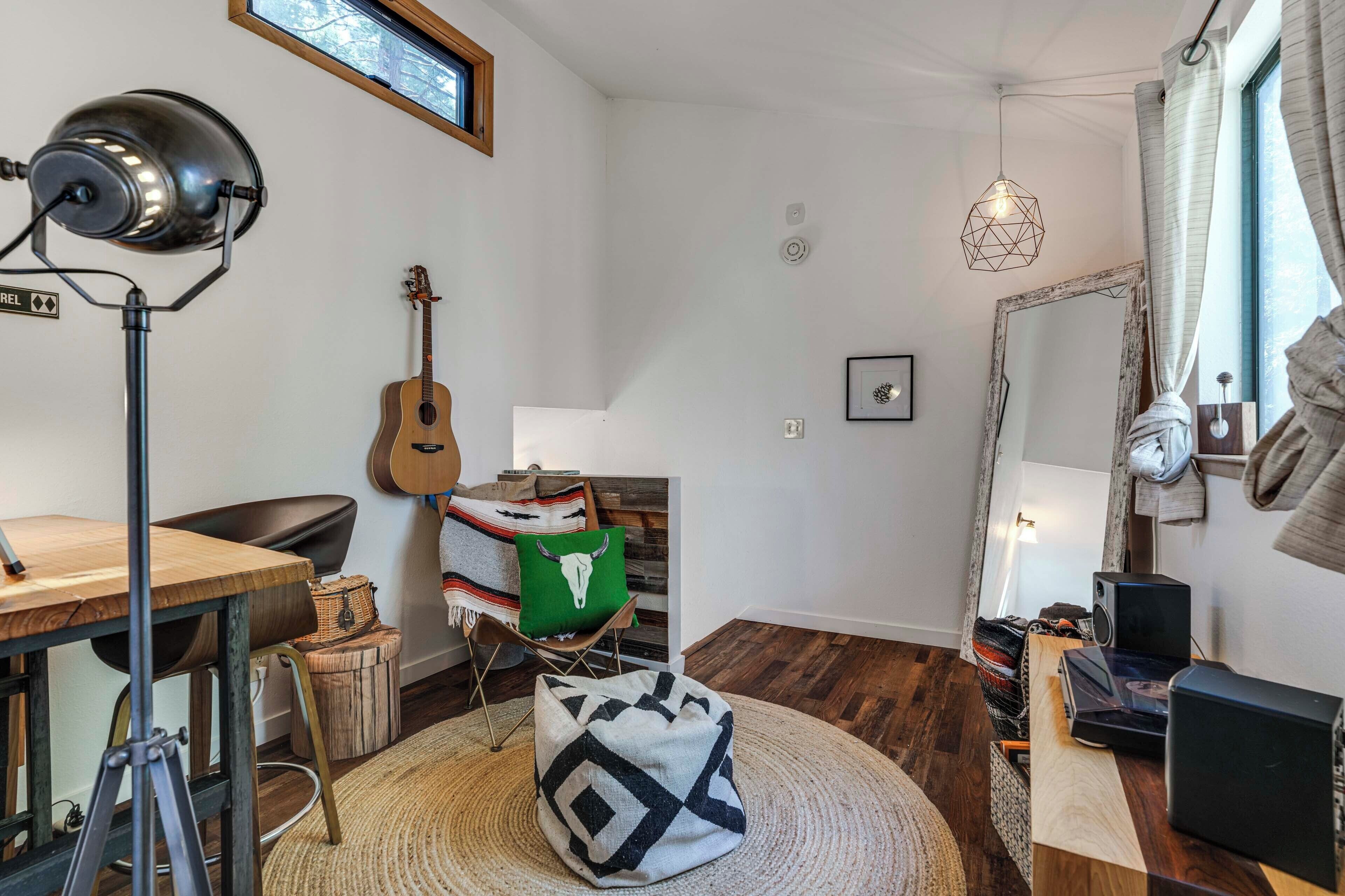 A room with a wooden desk, a guitar, a chair, a round rug, and a hanging light fixture.