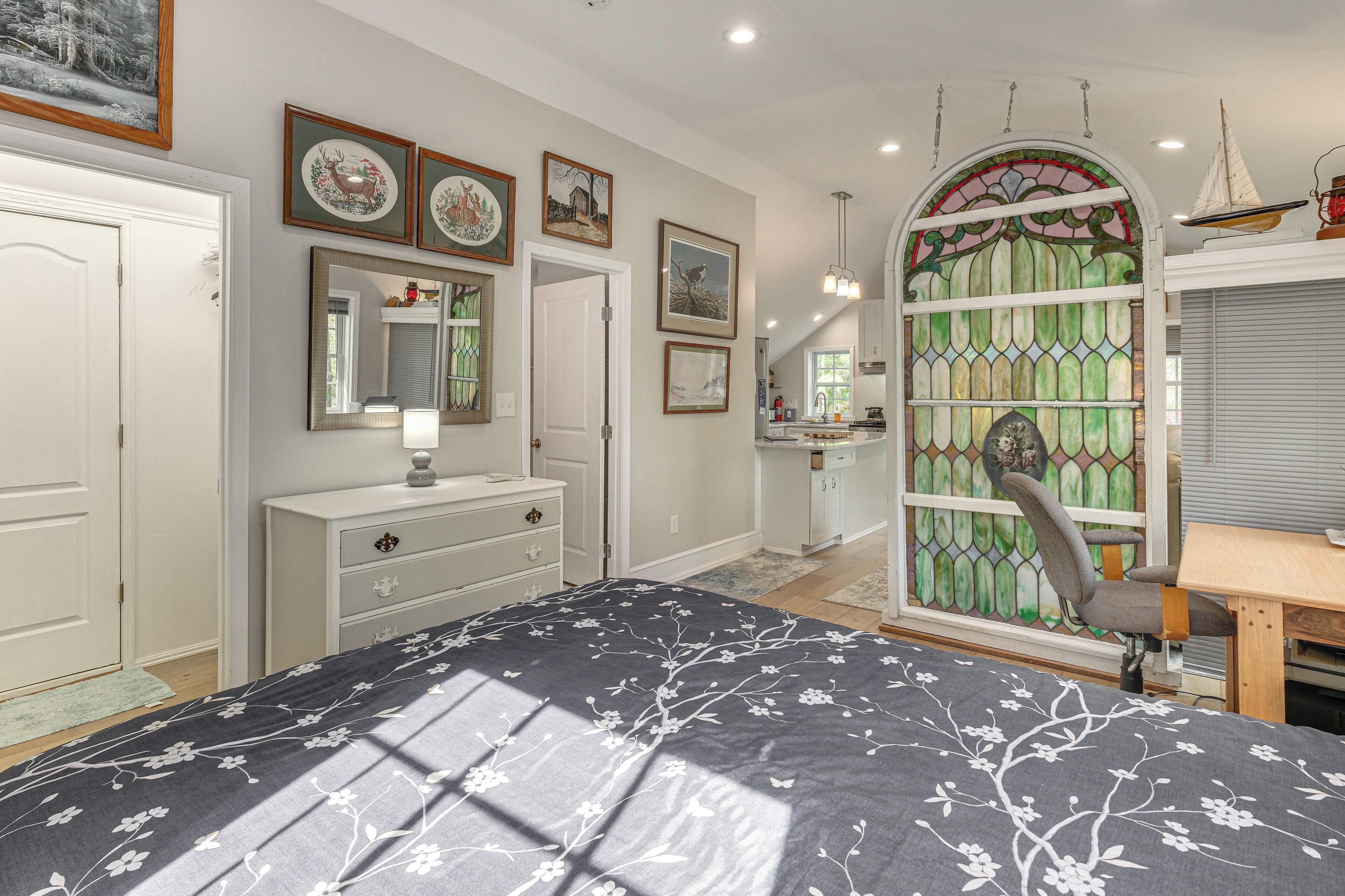 A bedroom with a bed, dresser, vanity, and a window with a stained glass design.