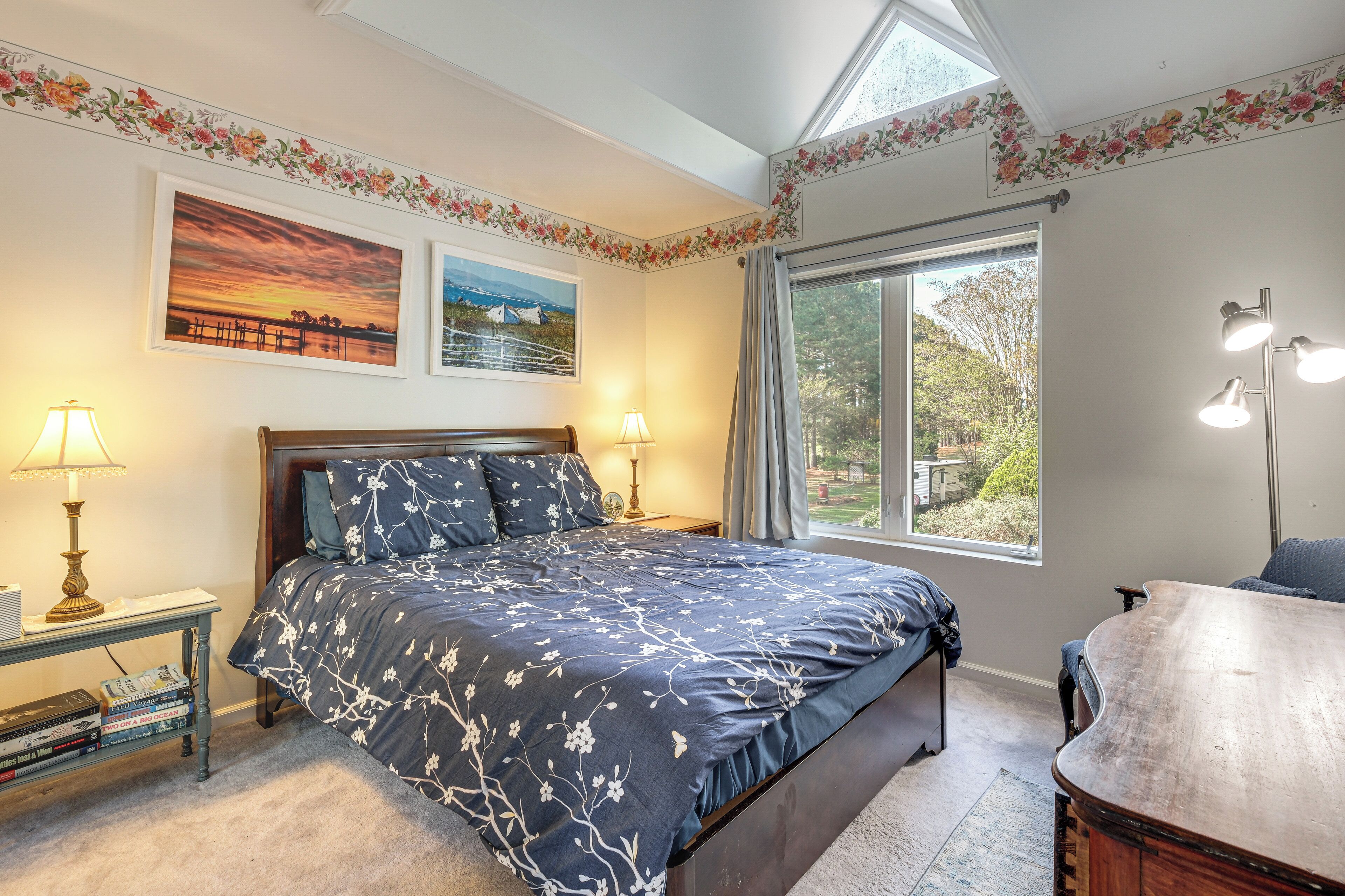 Ein Schlafzimmer mit einem Bett, zwei Nachttischlampen, einem Nachttisch mit Büchern, einem Fenster mit Blick und einer Decke mit Blumenverzierungen.