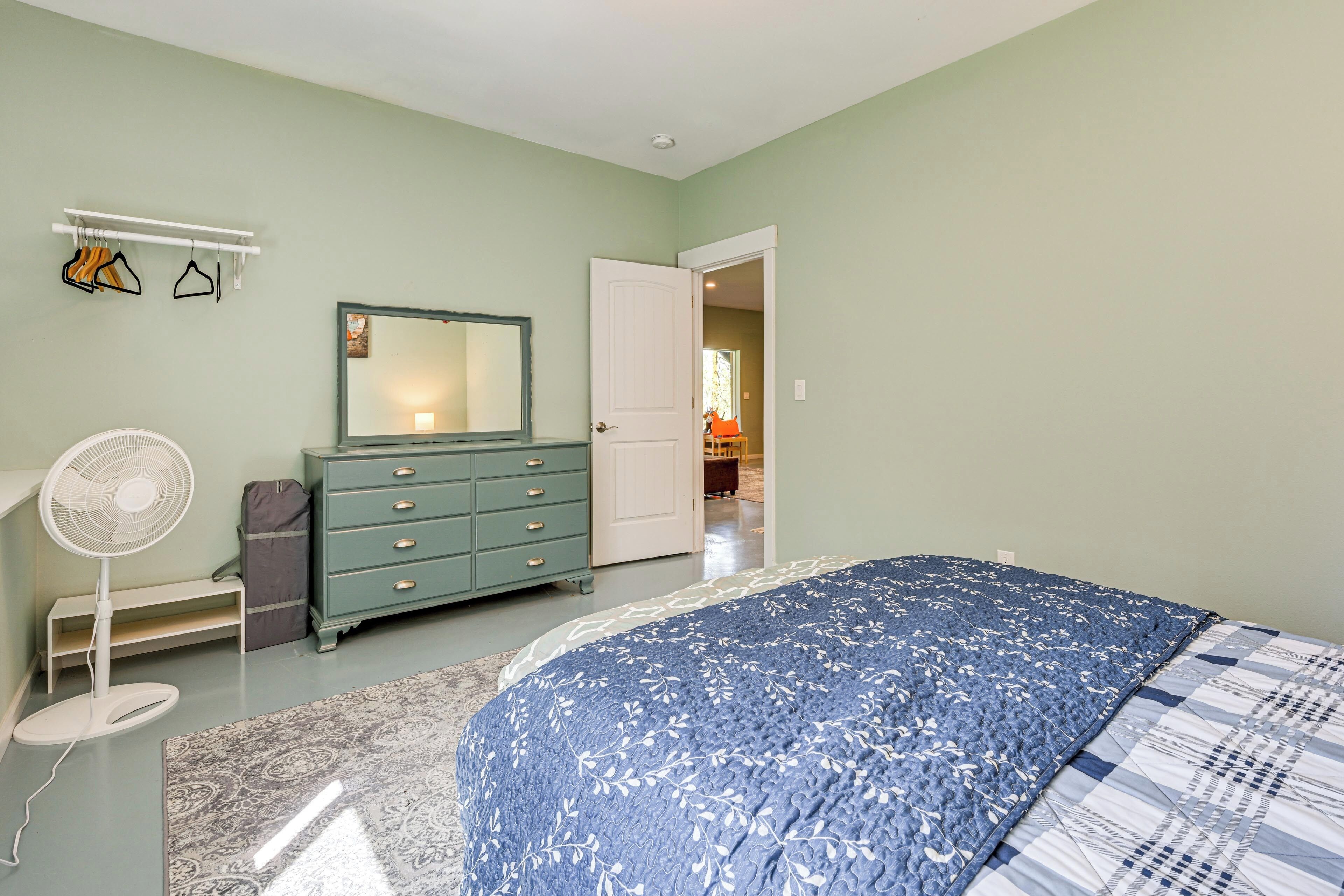 Ein Schlafzimmer mit Bett, Kommode, Spiegel, Ventilator und einer Tür zu einem weiteren Raum.