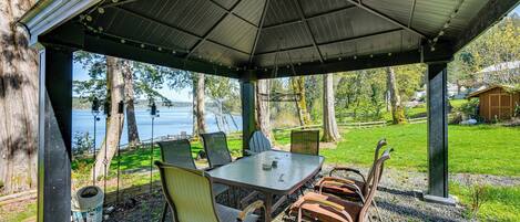 A covered outdoor seating area with a table and chairs, overlooking a body of water.