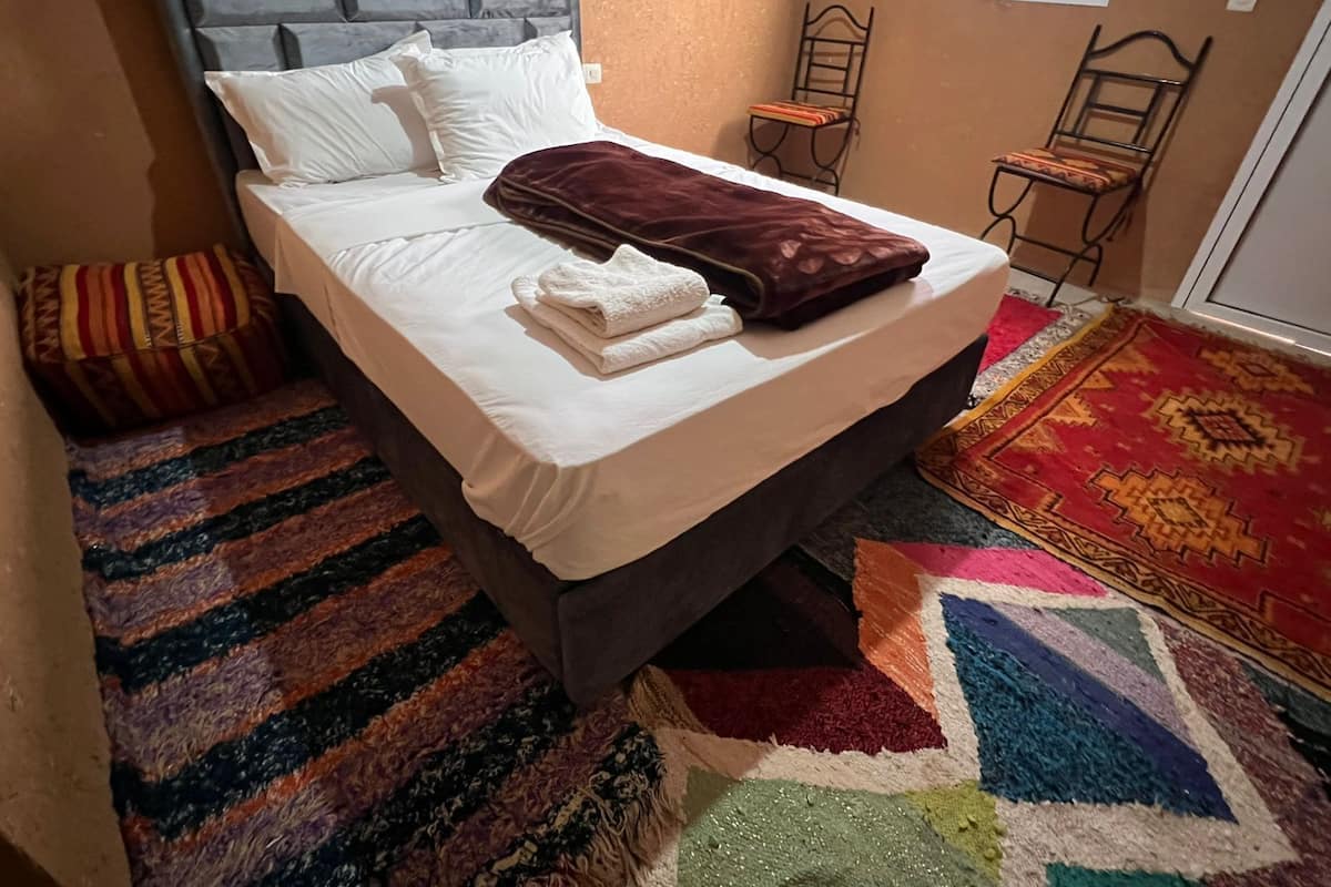 Una cama con cabecera, almohadas, una colcha y una toalla doblada. Hay dos sillas y una alfombra estampada.