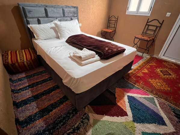A bed with a headboard, pillows, a blanket, and a folded towel. There are two chairs and a patterned rug.