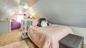 Ein gemütliches Schlafzimmer im Dachgeschoss mit schrägen Wänden, einer rosa Bettdecke, einem grauen Pouf und einem weißen, an der Wand befestigten Regal.