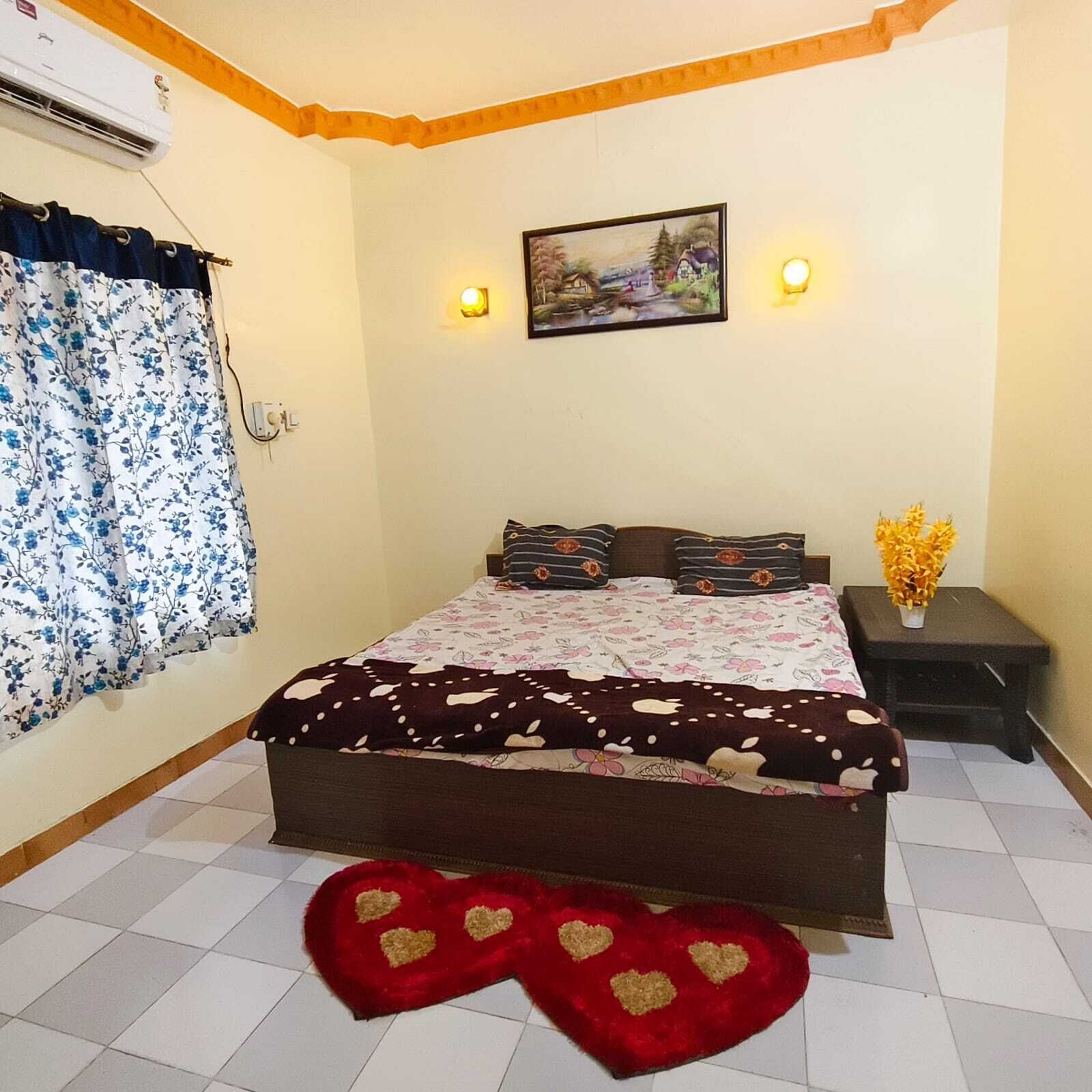 Un dormitorio con cama, mesita de noche, aire acondicionado montado en la pared, ventana con cortinas florales y un cuadro en la pared.