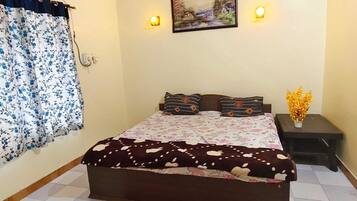 Un dormitorio con cama, mesita de noche, aire acondicionado montado en la pared, ventana con cortinas florales y un cuadro en la pared.