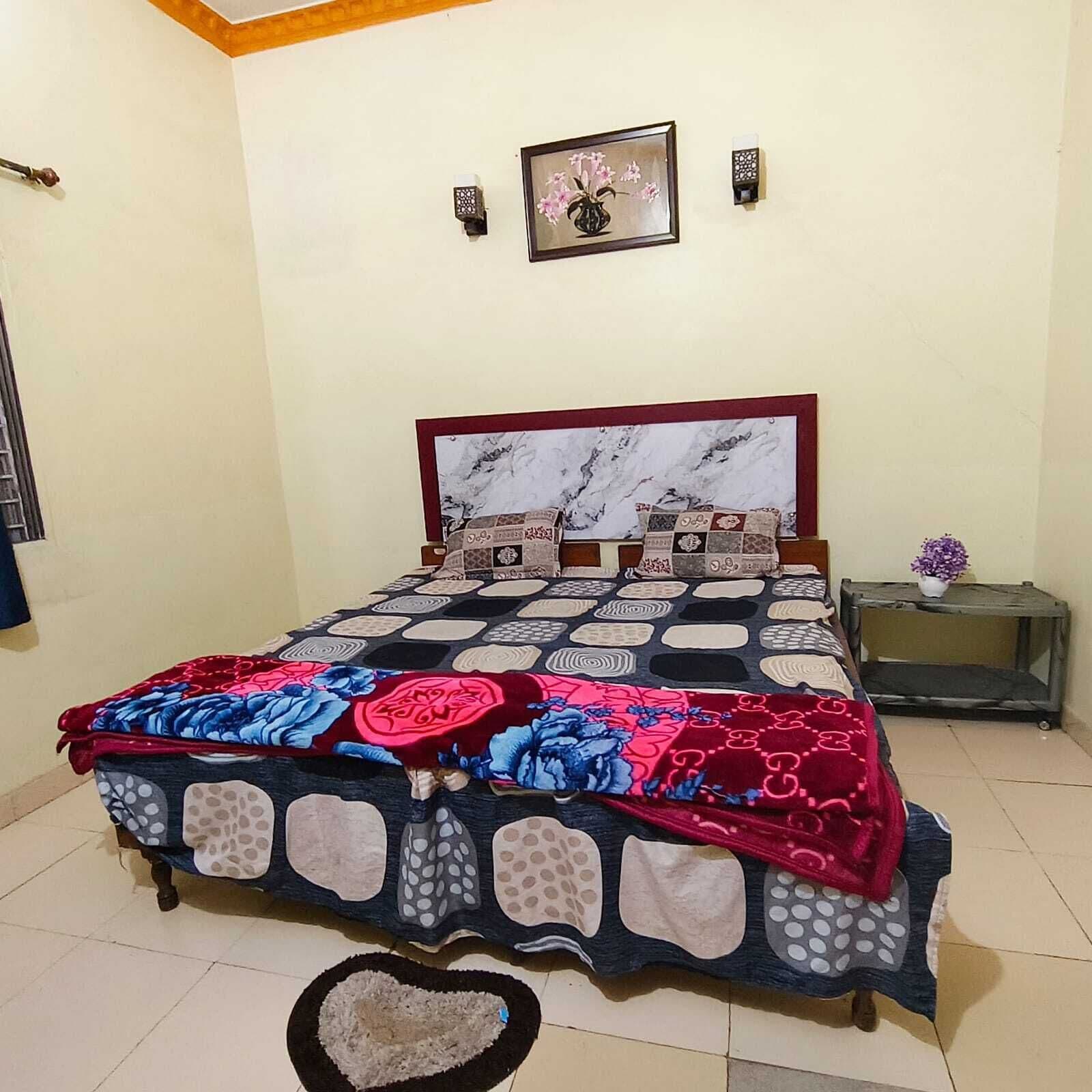 A bed with a patterned bedspread, a marble headboard, a small bedside table with a vase, and a wall-mounted picture.