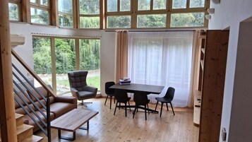 A room with a staircase, a dining table with chairs, a sofa, and a window with a view of greenery.