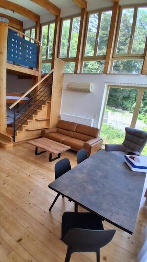 A wooden interior with a sofa, table, chairs, a staircase, and large windows.