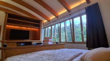 A bedroom with a large bed, a television on a wooden shelf, a window with curtains, and a view of trees.