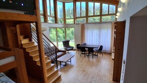 A wooden staircase leading to a dining area with a table and chairs, large windows, and a view of greenery outside.