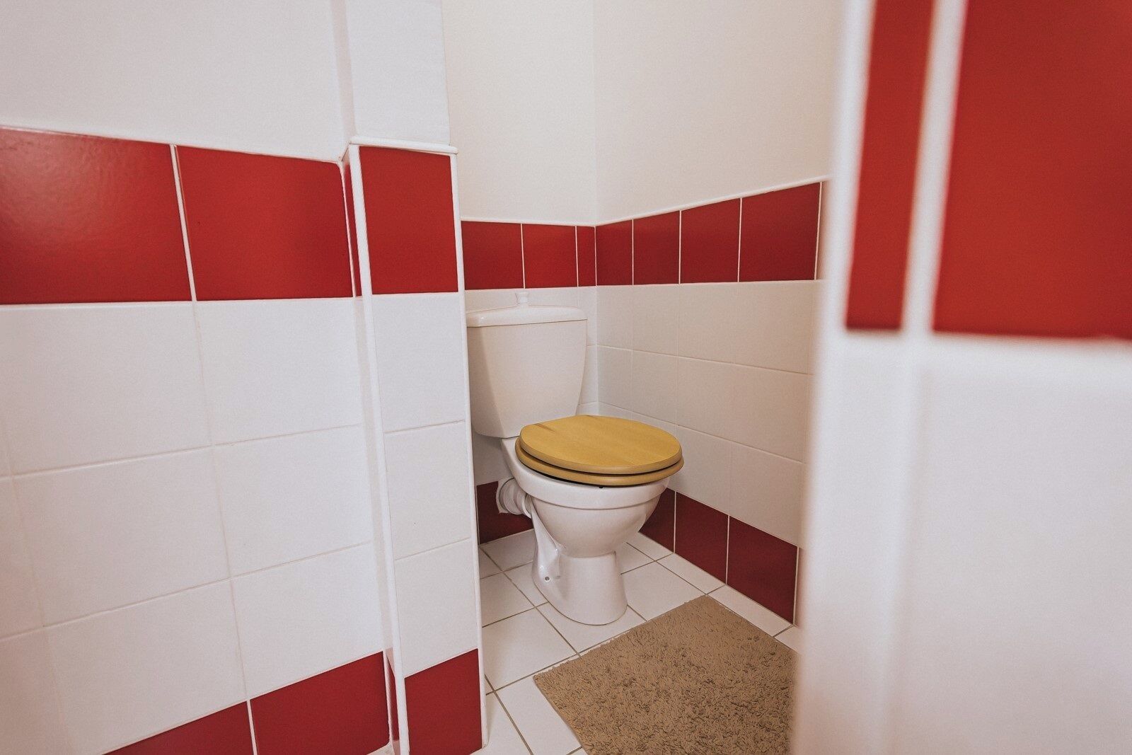A bathroom with white and red tiles, a toilet with a wooden seat, and a beige rug on the floor.