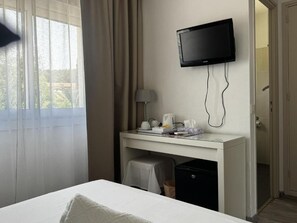 A hotel room with a bed, a desk with a television, a lamp, and a small trash bin. - Hôtel Le golfe bleu (Cavalaire-sur-Mer)