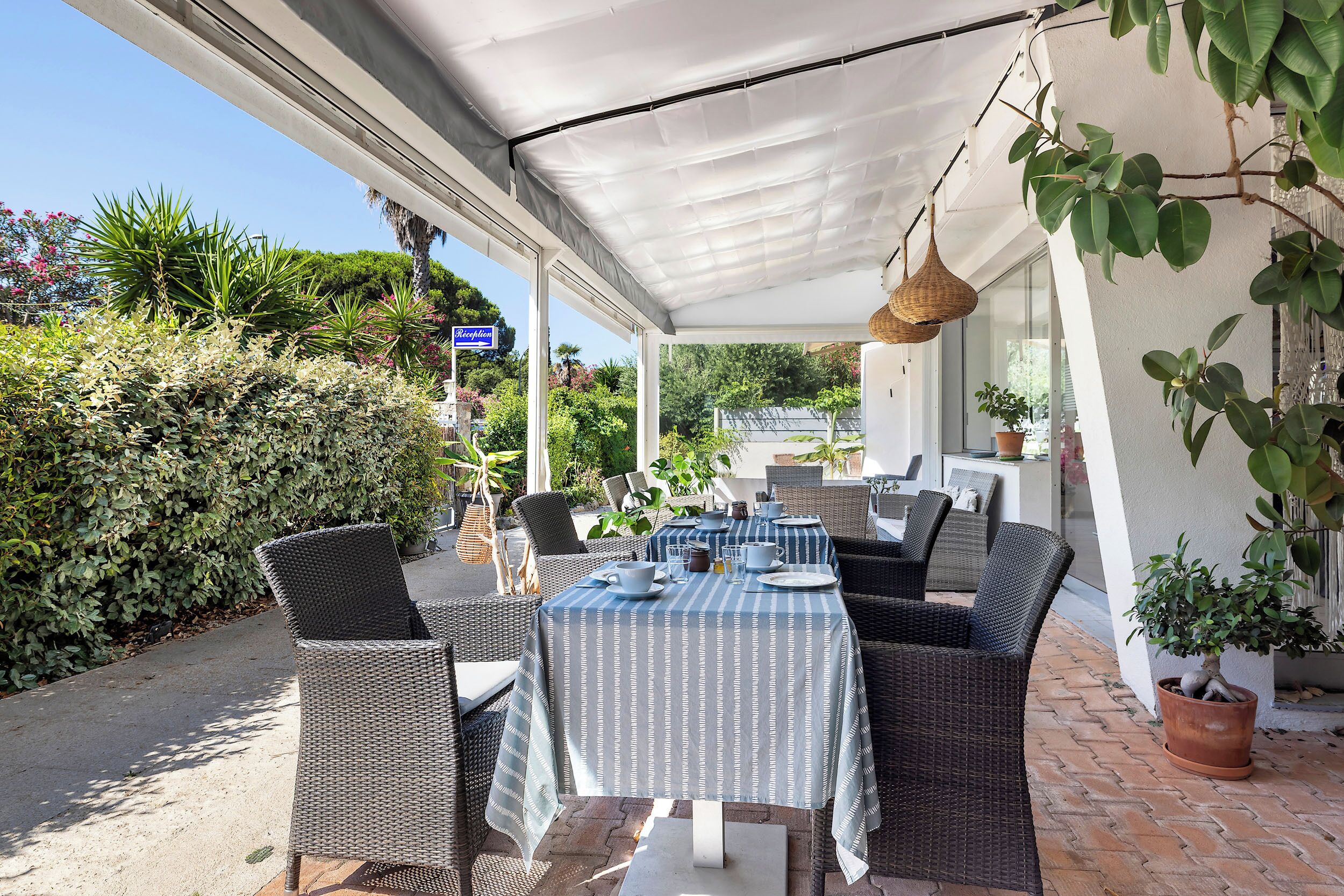 Une terrasse extérieure avec des meubles en osier, une table recouverte d’une nappe à rayures et des plantes en pot.
