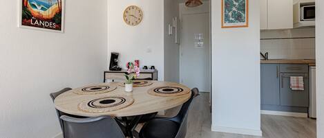 Un coin repas avec une table ronde, des chaises, une horloge et une cuisine en arrière-plan.