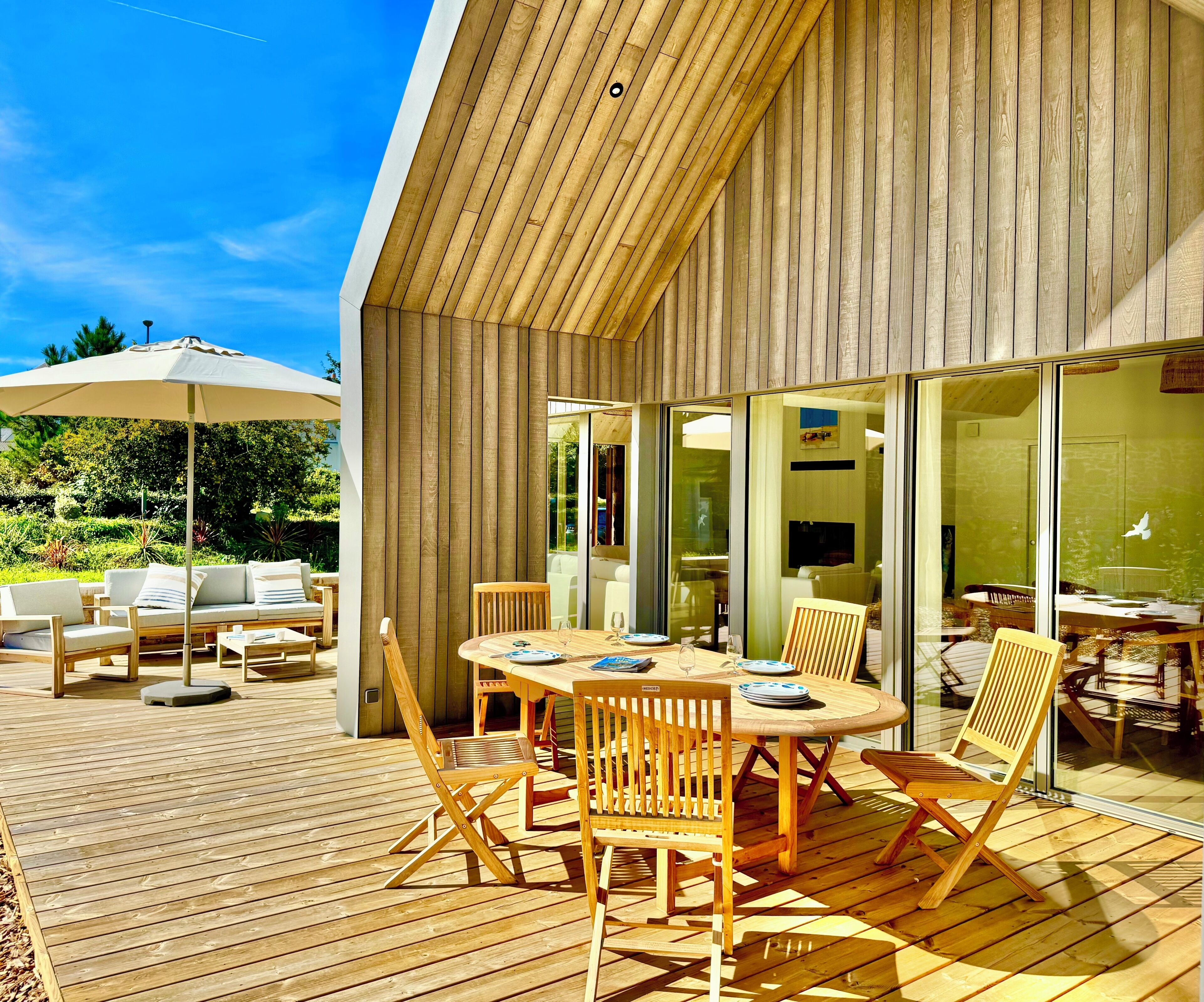 Un espacio moderno para comer al aire libre con muebles de madera, un gran paraguas y vistas del interior a través de puertas de cristal.