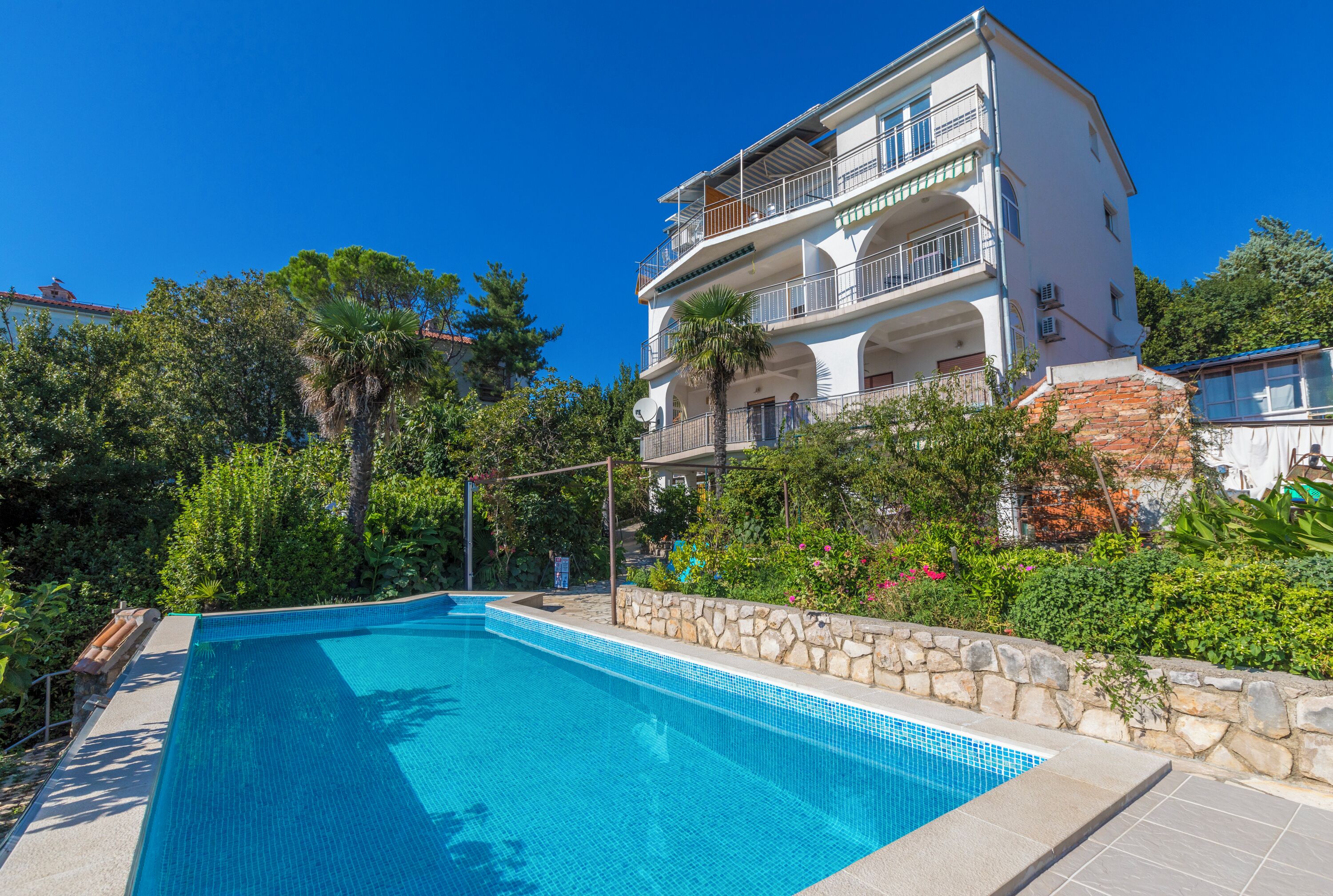 Um edifício com vários andares, piscina, vegetação exuberante e céu azul límpido.