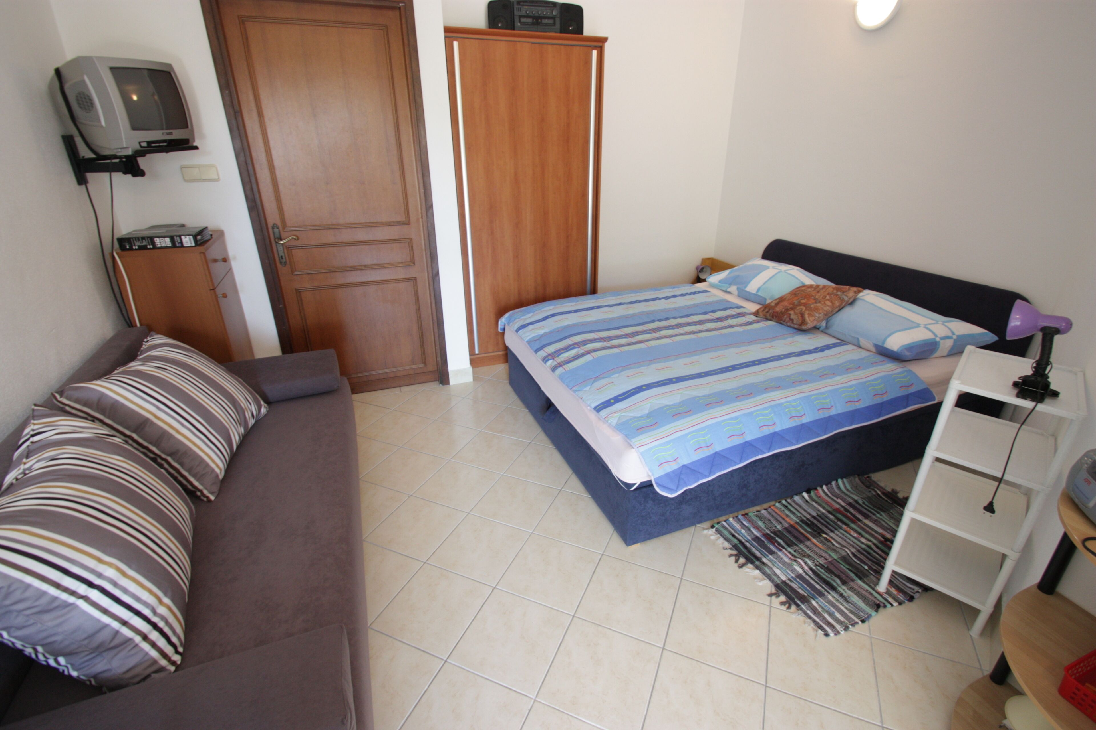 Quarto com cama, sofá, televisão, guarda-roupa e secretária com candeeiro.