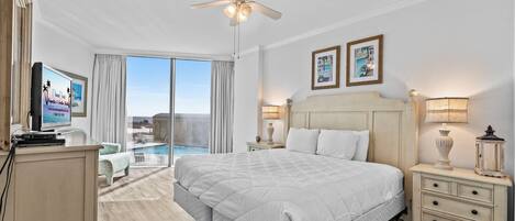 A hotel room with a large bed, a ceiling fan, a TV, a nightstand, a lamp, and a view of the pool.