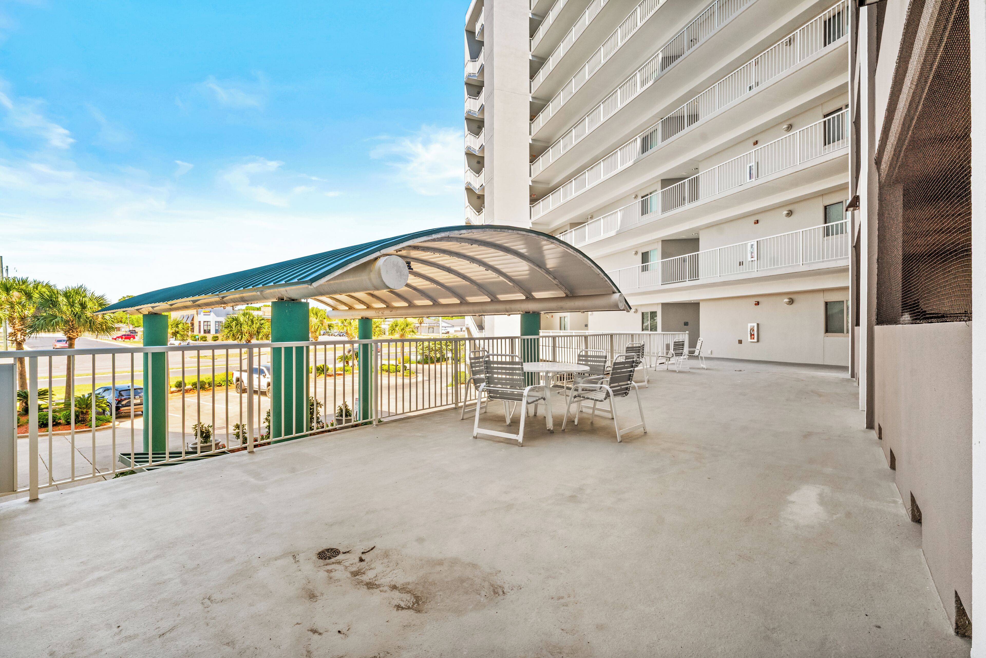 A covered outdoor seating area with tables and chairs, a parking lot, and a multi-story building with balconies.