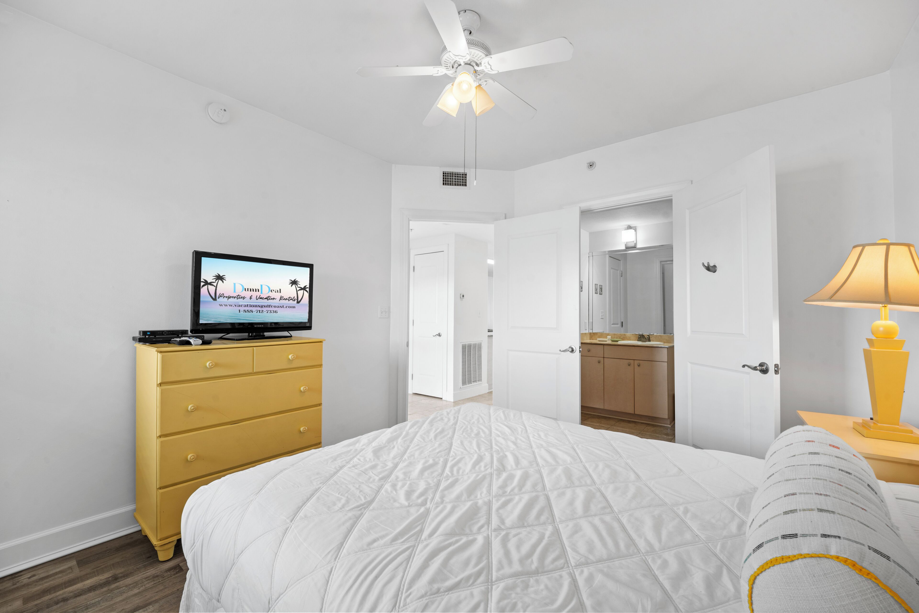 A bedroom with a bed, a TV on a dresser, a ceiling fan, and an open door leading to a bathroom.