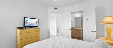 A bedroom with a bed, a TV on a dresser, a ceiling fan, and an open door leading to a bathroom.