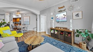 Een moderne woonkamer met een eethoek, een keuken op de achtergrond en een televisie waarop een advertentie met een strandthema wordt getoond.