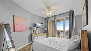 Een slaapkamer met een bed, een televisie, een dressoir, een plafondventilator en een groot raam.