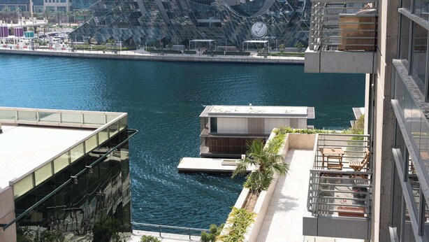 A modern building with a glass facade overlooking a body of water, a dock, and a small boat. - StayShort - Elegant 1BR in Prime Business Bay (Business Bay)