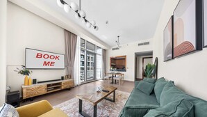 A modern living room with a sofa, coffee table, and a TV screen displaying 'BOOK ME'. - StayShort - Elegant 1BR in Prime Business Bay (Business Bay)